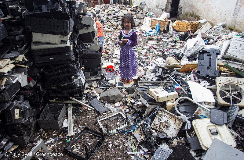Small girl in purple dress standing amidst endless piles of broken and discarded technology. 